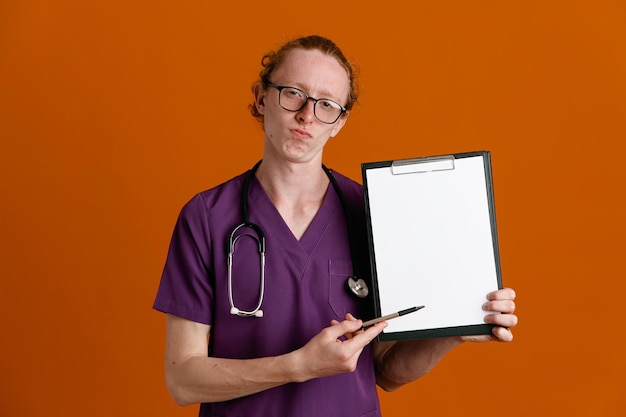 Tenuta sicura e punti con la penna negli appunti giovane medico maschio che indossa l'uniforme con uno stetoscopio isolato su sfondo arancione