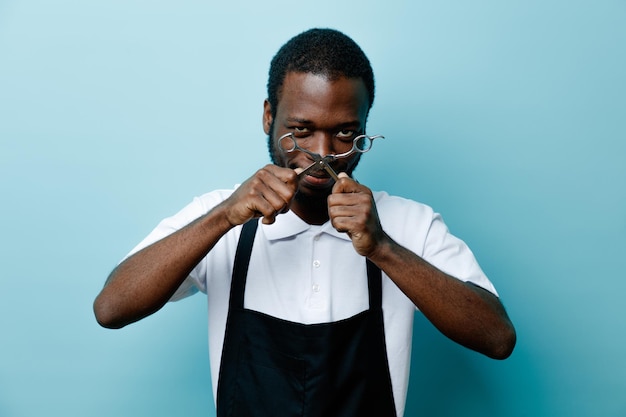 Confident holding scissors young african american barber in uniform isolated on blue background