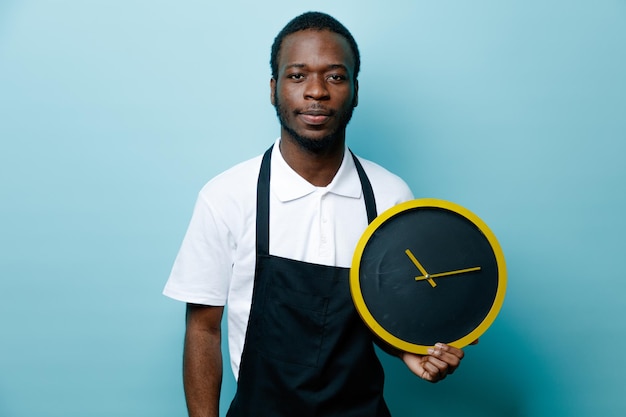 Confident holding wall clock young african american barber in uniform isolated on blue background