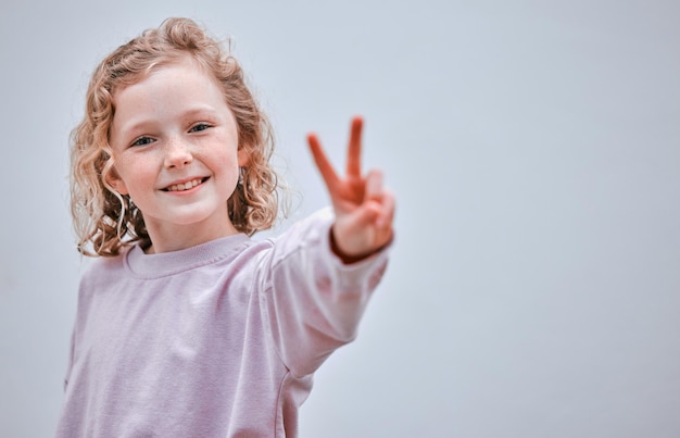 Уверенный в себе ребенок - счастливый ребенок Студийный снимок маленькой девочки, делающей знак мира на сером фоне