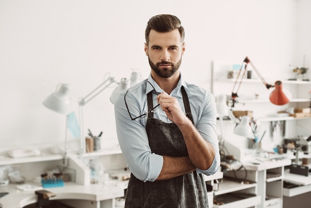 Confident leader. Portrait of confident male jeweler wearing apron and holding glasses while standing in jewelry making studio.