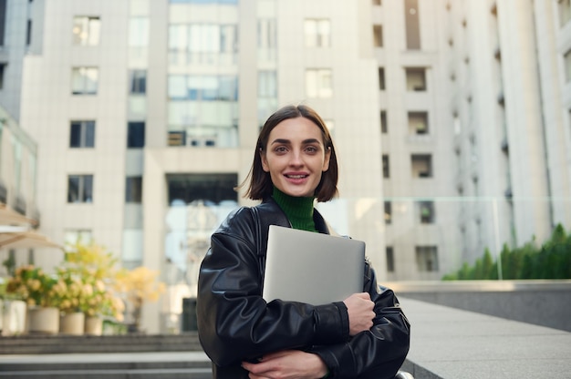 Confident lifestyle portrait of cheerful freelance woman with laptop, copywriter, motivated business person smiling looking confidently at camera in modern corporate high-rise buildings background