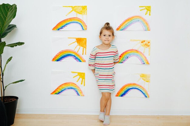 Confident little girl in a dress shirt next to six paintings of sun and rainbow