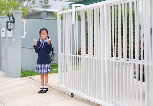 Уверенная маленькая девочка в школьной форме детского сада поднимает руки вверх перед уходом из дома