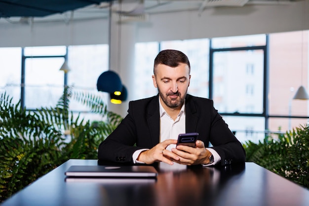 Confident male businessman in a business suit sits at a table with a phone and works remotely