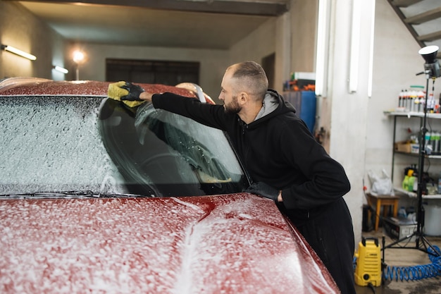 Confident male car wash worker cleaning the windshield of red luxury car with help of special foem