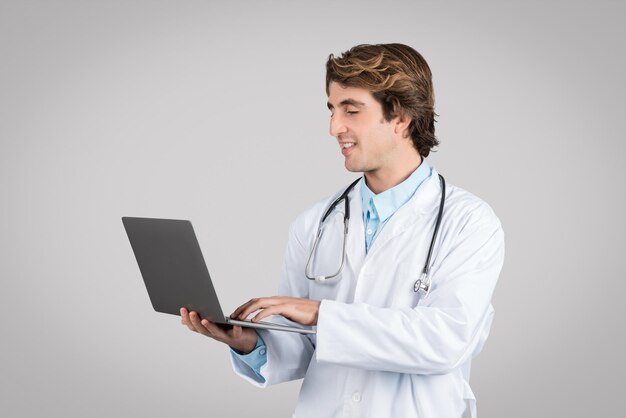 Уверенный в себе врач-мужчина в белом пальто со стетоскопом, пишущий на ноутбуке, изолированный на сером фоне
