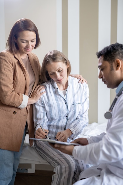 Fiducioso medico maschio in camice bianco che tiene un documento medico mentre la sua paziente mette la sua firma e la sua amica in piedi vicino