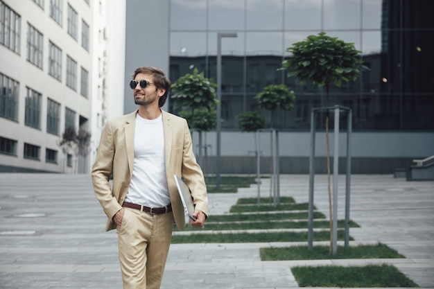 Confident male entrepreneur in elegant suit and sunglasses holding hand in pocket and wireless laptop