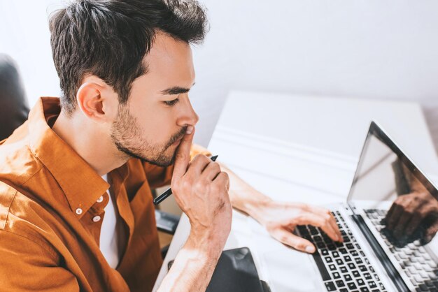 Confident male freelancer sitting in front of the computer with serious and thoughtful expression Young businessman in shirt with finger on mouth using pen and laptop for work sitting in the office