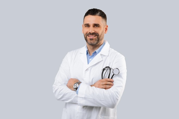 Foto fiducioso medico maschio in camice bianco che tiene in mano lo stetoscopio in posa con le braccia conserte