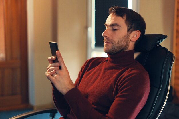 Un uomo fiducioso controlla le notizie al telefono mentre è seduto su una sedia in pelle in ufficio