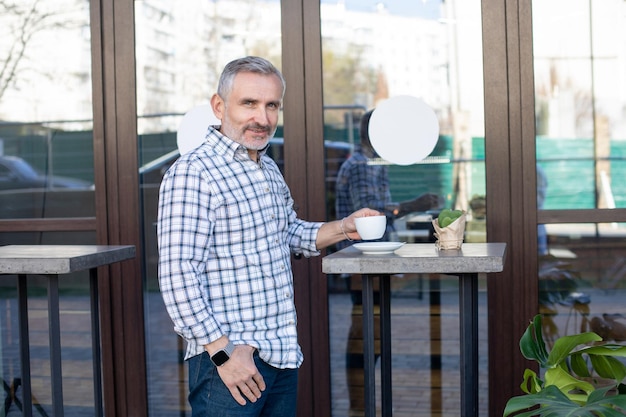 Confident man in plaid shirt standing near the table in a street cafe