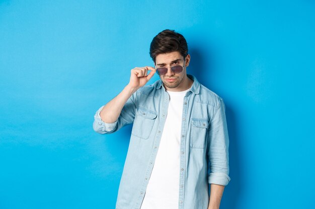 Confident man put on sunglasses, looking cool and sassy, standing over blue wall