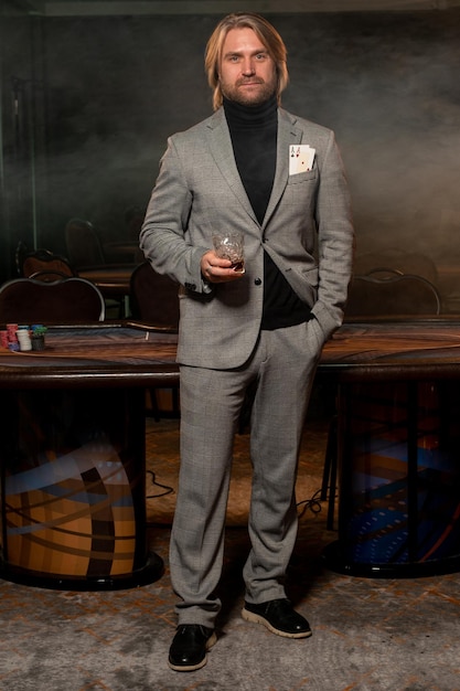 Фото Уверенный в себе мужчина с парой тузов в кармане куртки и стаканом напитка в руке стоит в казино