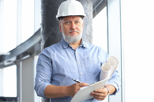 Photo confident mature business man in hardhat holding blueprint while standing indoors.