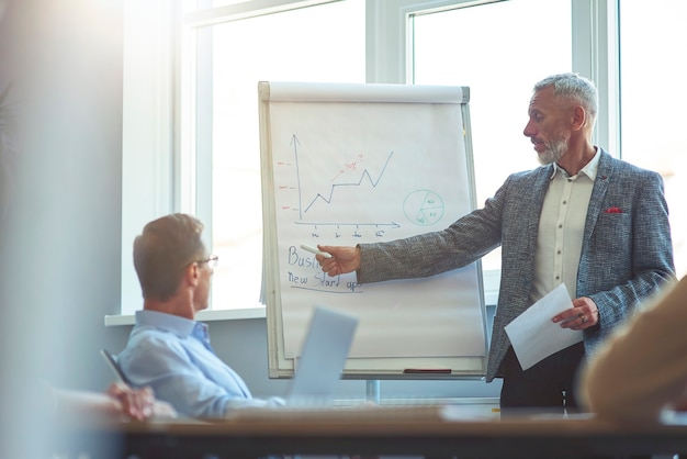 Photo confident mature businessman pointing at flip chart and explaining something to colleagues while