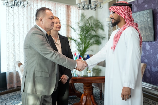 Confident mature businessman in suit greeting foreign partner by handshake and looking at him after negotiating and making deal