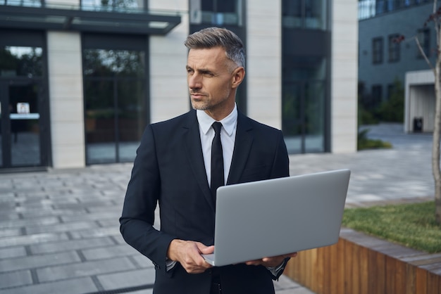 Confident mature businessman working on laptop while standing outdoors near office building