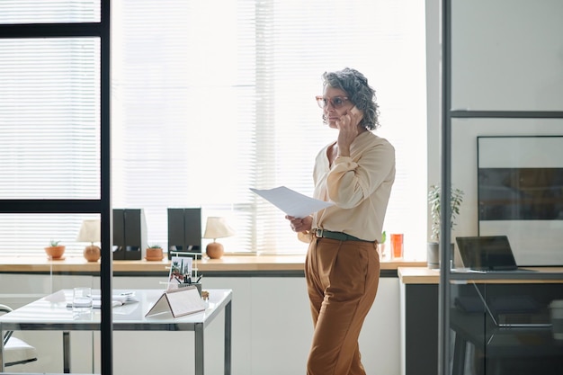 confident mature businesswoman with cellphone by ear standing in office
