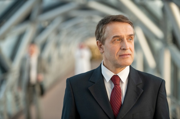 Confident mature delegate or businessman in elegant suit standing in front of camera against long aisle inside contemporary business center