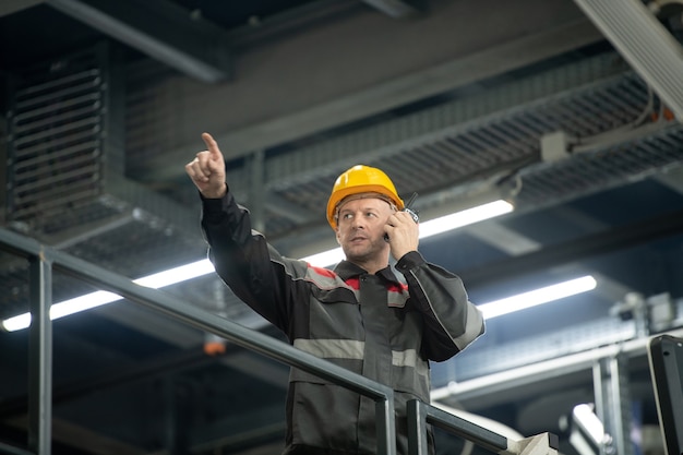 Confident mature foreman with hardhat on head and walkie talkie by his ear pointing forwards during work in large polymer processing factory