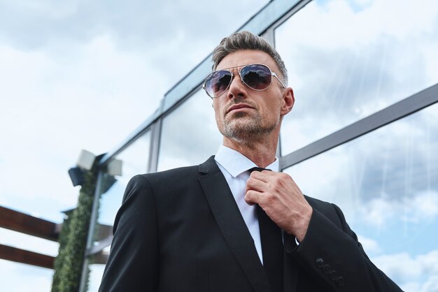 Confident mature man carrying laptop and adjusting necktie while walking on rooftop terrace