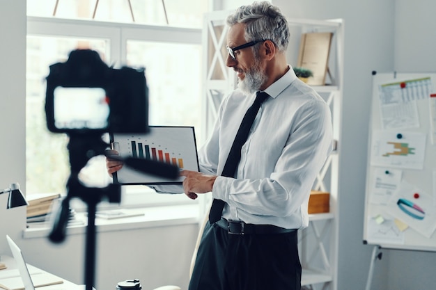 Fiducioso uomo maturo in elegante camicia e cravatta che mostra il grafico e condivide l'esperienza aziendale mentre realizza video sui social media