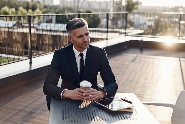 Confident mature man in formalwear enjoying coffee while sitting in cafe outdoors