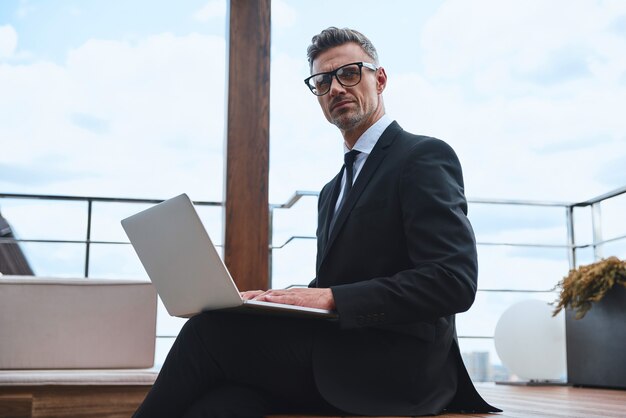 Confident mature man working on laptop while standing on the rooftop terrace