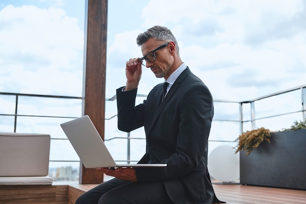 Confident mature man working on laptop while standing on the rooftop terrace