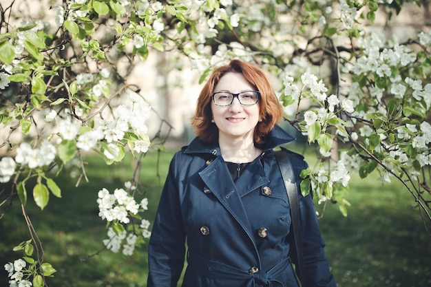 Confident middle aged Armenian woman in a blue trench coat and glasses under the blooming tree Smiling looking at the camera