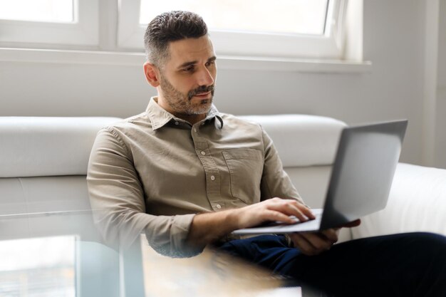自信を持って中年の実業家がオフィスのソファに座って、新しいノートパソコンで作業しています
