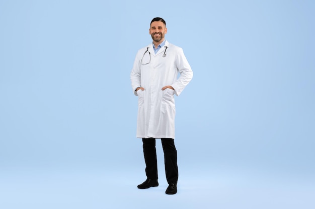 Photo confident middle aged male doctor in white coat posing with hands in pockets on blue background full length