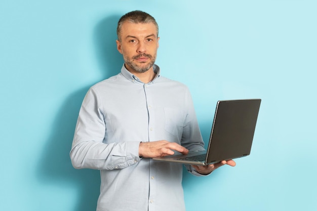 Confident middle aged man holding and using laptop computer standing over blue studio background