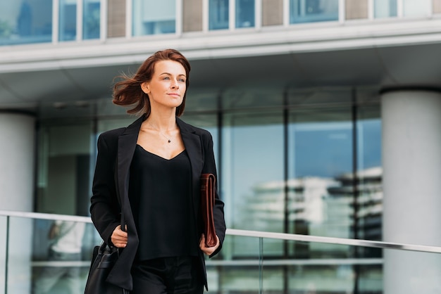 Уверенная в себе деловая женщина среднего возраста, держащая кожаную папку, идущая против офисного здания