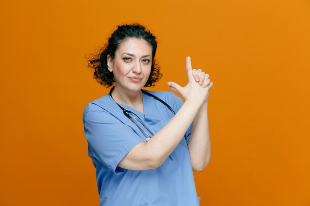 Fiduciosa dottoressa di mezza età che indossa uniforme e stetoscopio intorno al collo guardando la fotocamera che mostra il gesto della pistola isolato su sfondo arancione