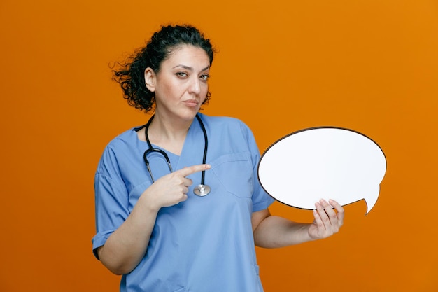 Confident middleaged female doctor wearing uniform and stethoscope around her neck showing chat bubble pointing at it looking at camera isolated on orange background