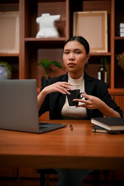 Confident millennial Asian businesswoman holding a cup of coffee sitting at her office desk
