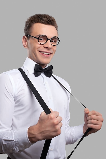 Confident nerd. Cheerful young man in bow tie adjusting his suspenders and smiling at camera while standing against grey background