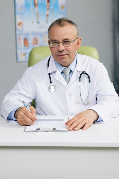 Confident old mature male head doctor physician in white medical uniform in glasses sitting at workplace.