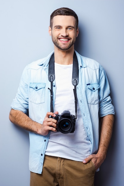 Confident photographer. Handsome young man holding digital camera and smiling while standing against grey background