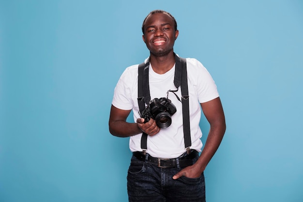 Confident photographer having professional camera smiling heartily while standing on blue background. Handsome looking young man wearing fashion clothes while having photo device.