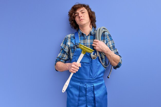 Confident plumber man with curly hair in uniform holding toilet plunger isolated on blue studio background