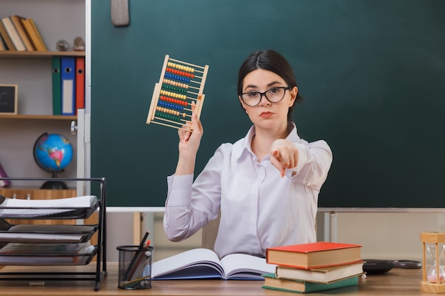 Punti fiduciosi alla telecamera giovane insegnante femminile che indossa occhiali con abaco seduto alla scrivania con strumenti scolastici in classe