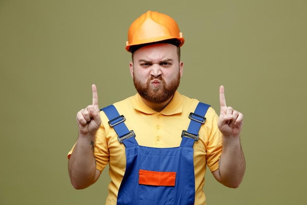 Photo confident points at up young builder man in uniform isolated on green background