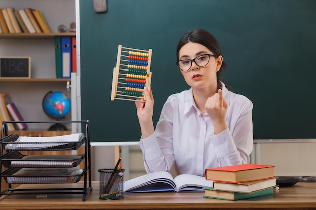 confident points at up young female teacher wearing glasses holding abacus sitting at desk with school tools in classroom