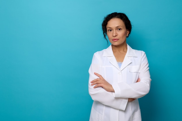 Fiducioso ritratto di bella dottoressa serena in abito medico bianco, guardando la telecamera in posa con le braccia incrociate su sfondo blu colorato con copia spazio per la pubblicità medica