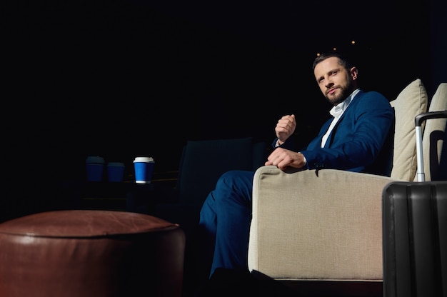 Confident portrait of a handsome attractive young man in a business suit sits in an armchair in the lounge area of a luxury hotel during a business trip and looks confidently to the side cute smiling