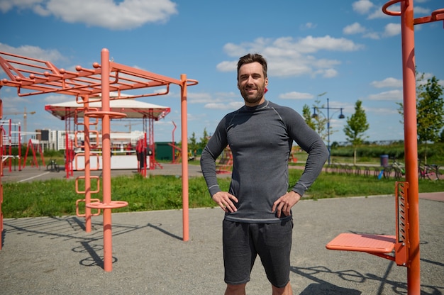 Confident portrait of a handsome Caucasian sportsman athlete standing on the sports field with arms on waist, smiling looking at the camera. Handsome Caucasian guy in sportswear resting after workout